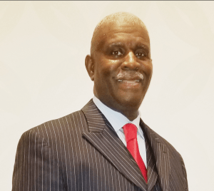 Michael Bart Mathews, smiling in a formal suit with a pinstripe pattern and a red tie, representing entrepreneurship and leadership in the Business Booster Today magazine context.